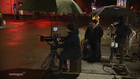 Ein Kamerateam sitzt bei Nacht unter Regenschirmen an einer Straße. © Screenshot 