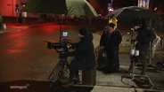Ein Kamerateam sitzt bei Nacht unter Regenschirmen an einer Straße. © Screenshot 