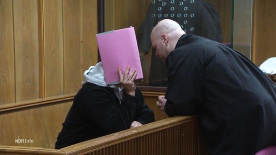 Eine Person in einem Gerichtssaal verdeckt sein Gesicht mit einer Mappe und sein Verteidiger spricht zu ihm. © Screenshot 