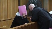 Eine Person in einem Gerichtssaal verdeckt sein Gesicht mit einer Mappe und sein Verteidiger spricht zu ihm. © Screenshot 