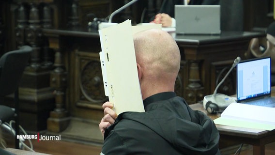 Ein junger Mann mit Glatze sitzt in einem Gerichtssaal, er hält sich eine Akte vors Gesicht. © Screenshot 