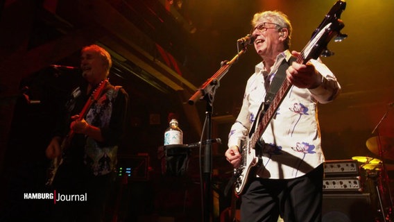 Der Bassist Graham Gouldman der Band 10cc singt auf der Bühne in ein Mikrophon, neben ihm steht ein weiteres Bandmitglied. © Screenshot 