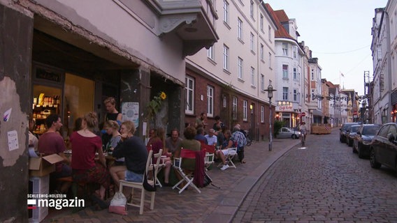Mehrere Menschen sitzen vor einem Café in einer kleinen Straße. © Screenshot 