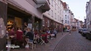 Mehrere Menschen sitzen vor einem Café in einer kleinen Straße. © Screenshot 