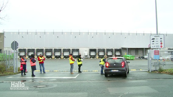Vor dem Zufahrtstor eines größeren Paketverteilungszentrums stehen mehrere Menschen in Warnwesten. © Screenshot 