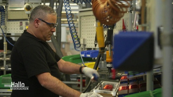 Ein Mann steht in einer Fabrikhalle an einer Werkbank mit Schutzbrille. © Screenshot 