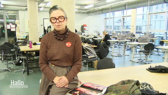 Modedesignerin Martina Glomb sitzt an der Kante eines Ateliertischs in einem größeren Raum. © Screenshot 