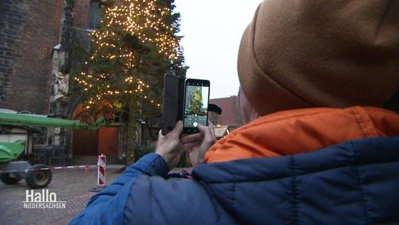 Eine Person in Winterjacke und mit Mütze nimmt mit dem Handy ein Foto von einem großen Weihnachtsbaum vor einer Kirche auf. © Screenshot 