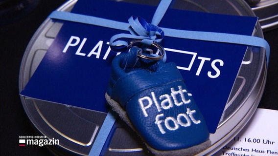 Nahaufnahme eines Kinderschuhs mit der Aufschrift "Plattfoot", platziert auf einer Filmrolle. © Screenshot 