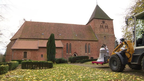 Ein Traktor mit Gabelfunktion fährt eine Kirchenglocke zu einer Kirche. © Screenshot 