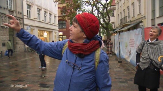 Eine Stadtführerin zeigt auf ein Gebäude während sie etwas in einer Fußgängerzone ihren Zuhörenden erklärt. © Screenshot 