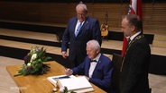 Manfred Tepper sitzt an einem Tisch. Neben ihm stehen zwei Männer. © Screenshot 