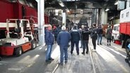 Mehrere Menschen stehen in einer Halle zwischen Eisenbahnen. © Screenshot 
