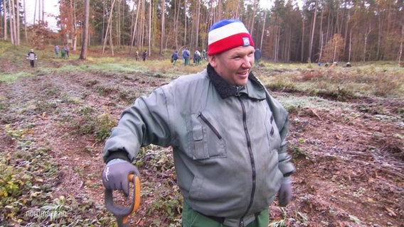 Ein Mann mit Mütze und Spaten auf einer Waldlichtung. © Screenshot 