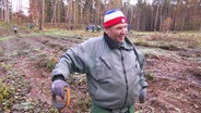 Ein Mann mit Mütze und Spaten auf einer Waldlichtung. © Screenshot 