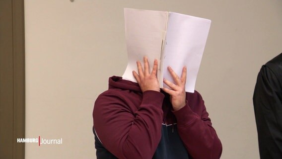 Eine Person hält sich in einem Gerichtssaal eine Akte vor das Gesicht. © Screenshot 