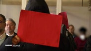 Einer der verurteilten Täter hält sich eine rote Mappe vor sein Gesicht. © Screenshot 