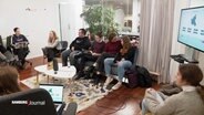 Menschen auf Sitzmöbeln und mit mobilen Endgeräten in einem Raum im Kreis sitzend. © Screenshot 