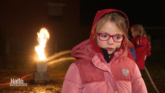 Ein Mädchen spricht in die Kamera und im Hintergrund wird ein Feuer gelöscht. © Screenshot 