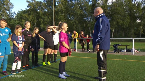 Ein Trainer steht auf einem Sportplatz vor einer Gruppe Kinder, ein Mädchen steht direkt vor ihm. © Screenshot 