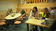 Verschiedene Personen sitzen um Tische und sprechen miteinander. Im Hintergrund hängt eine Fähnchenkette mit der Deutschlandflagge. © Screenshot 
