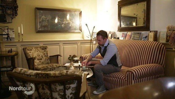 Ein Mann sitzt auf einer Couch und ttrinkt Kaffee in einem im Stil der 20er und 30er Jahre eingerichteten Raum. © Screenshot 