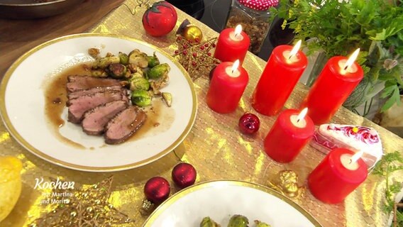 Ein mit roten Kerzen, goldener Tischdecke und Kugeln weihnachtlich gedeckter Tisch. Auf einem Teller mit Goldrand ist ein Braten angerichtet. © Screenshot 