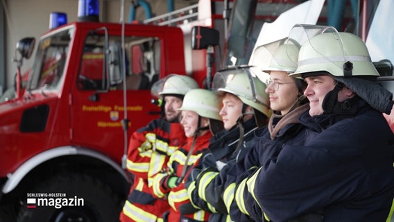 Schräg von der Seite zu sehen stehen fünf Personen in Feuerwehr-Arbeitskleidung in einer Reihe, im Hintergrund parkt ein Fahrzeug der Feuerwehr. © Screenshot 