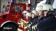 Schräg von der Seite zu sehen stehen fünf Personen in Feuerwehr-Arbeitskleidung in einer Reihe, im Hintergrund parkt ein Fahrzeug der Feuerwehr. © Screenshot 