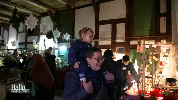 Ein Mann mit einem Kind auf den Schultern besucht eine Weihnachtsausstellung mit Kunsthandwerk. © Screenshot 