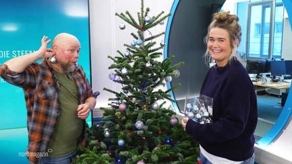 Theresa und ein mit Weihnachtsbaumkugeln behangener Stefan stehen im Studio. © Screenshot 