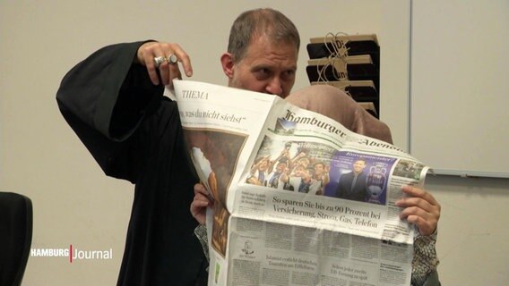 Im Gerichtssaal versteckt sich eine Frau neben ihrem Verteidiger hinter einer Zeitung. © Screenshot 