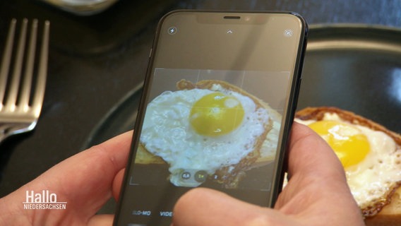 Zwei Hände halten ein Smartphone, auf dem Dispplay sieht man, wie ein Foto von einem Brot mit Spiegelei gemacht wird. Im Hintergrund sieht man den Teller mit dem Brot. © Screenshot 