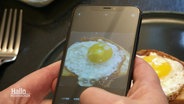 Zwei Hände halten ein Smartphone, auf dem Dispplay sieht man, wie ein Foto von einem Brot mit Spiegelei gemacht wird. Im Hintergrund sieht man den Teller mit dem Brot. © Screenshot 