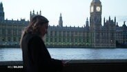 Eine junge Frau steht an der Themse – im Hintergrund ist das Londoner Regierungsviertel zu sehen. © Screenshot 