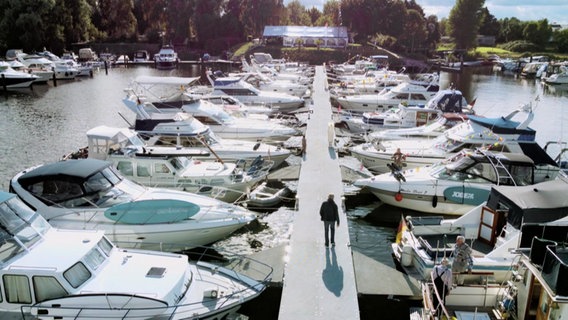 Aus der Vogelperspektive: Eine Person geht einen Steg an einem Hafen entlang, links und rechts sind Motorboote festgemacht. © Screenshot 