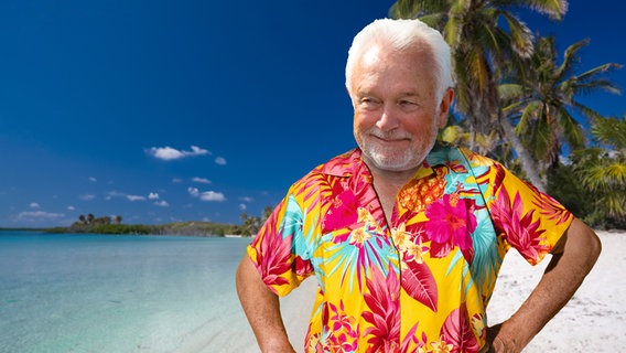 Wolfgang Kubicki in einem Hawaii-Hemd an einem Strand mit Palmen. (extra 3 vom 06.12.2023 im NDR Fernsehen) © NDR 