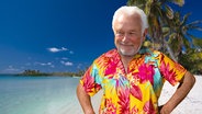 Wolfgang Kubicki in einem Hawaii-Hemd an einem Strand mit Palmen. (extra 3 vom 06.12.2023 im NDR Fernsehen) © NDR 