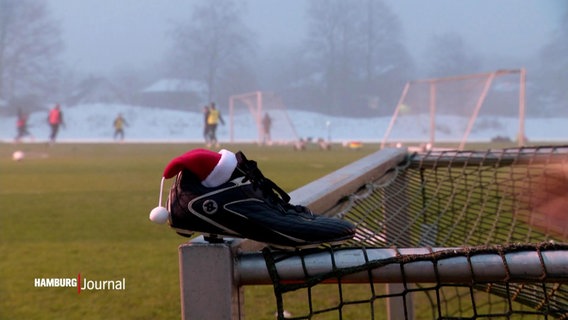 Ein Stollenschuh mit einer Nikolausmütze auf einem Torpfosten. Im Hintergrund das Training der Herren-Mannschaft des FC St. Pauli. © Screenshot 