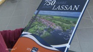 Eine Festschrift mit dem Titel "750 Jahre Stadt Lassan" wird von einer Person gehalten. © Screenshot 