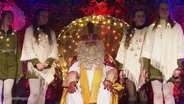 Der Nikolaus sitzt auf einer Bühne. Seine Helferinnen stehen links und rechts von ihm. © Screenshot 