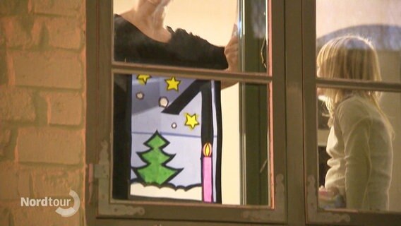 Eine Frau klebt ein Fensterbild auf die Scheibe. Darauf steht eine große Eins mit einer Kerze und einem Tannenbaum. © Screenshot 