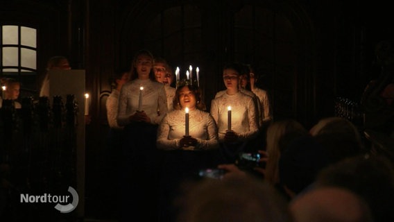 Eine Gruppe von weiß gekleideten Jugendlichen erhellen die Dunkelheit mit Kerzenschein. © Screenshot 