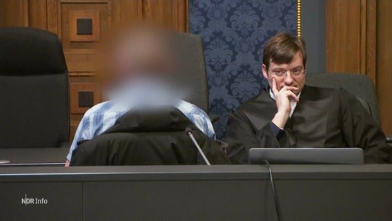 Der Angeklagte sitzt in einem Gerichtssaal, sein Kopf wurde unkenntlich verpixelt. Neben ihm sitzt ein Staatsanwalt und schaut ihn an. © Screenshot 