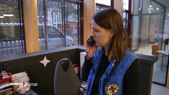 Eine junge Frau spricht in ein Telefon. Sie trägt eine Weste der Bahnhofsmission. © Screenshot 