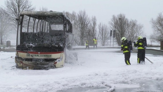 Zwei Feuerwehrleute stehen in voller Sicherheitskleidung in einer verschneiten Landschaft neben den ausgebrannten Überresten eines Reisebusses. © Screenshot 