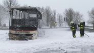 Zwei Feuerwehrleute stehen in voller Sicherheitskleidung in einer verschneiten Landschaft neben den ausgebrannten Überresten eines Reisebusses. © Screenshot 
