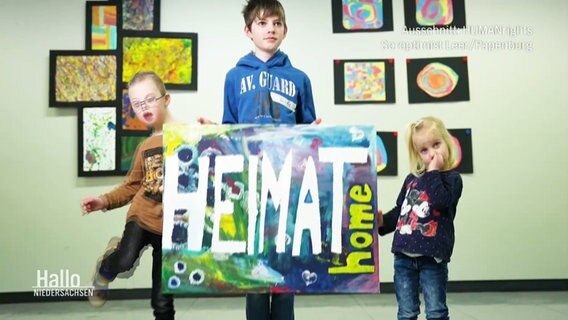 Drei Kinder halten eine Tafel mit der Aufschrift "Heimat - home" © Screenshot 