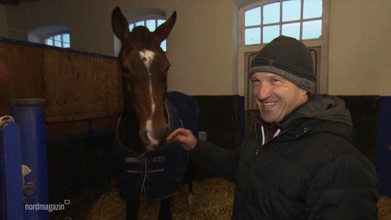 Springreiter Holger Wulschner steht in einem Stall neben einem Pferd und lächelt. © Screenshot 