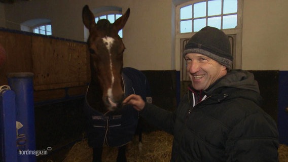 Springreiter Holger Wulschner steht in einem Stall neben einem Pferd und lächelt. © Screenshot 